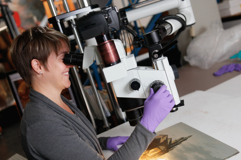 Woman examining oil painting under a microscope in an art conservation laboratory.